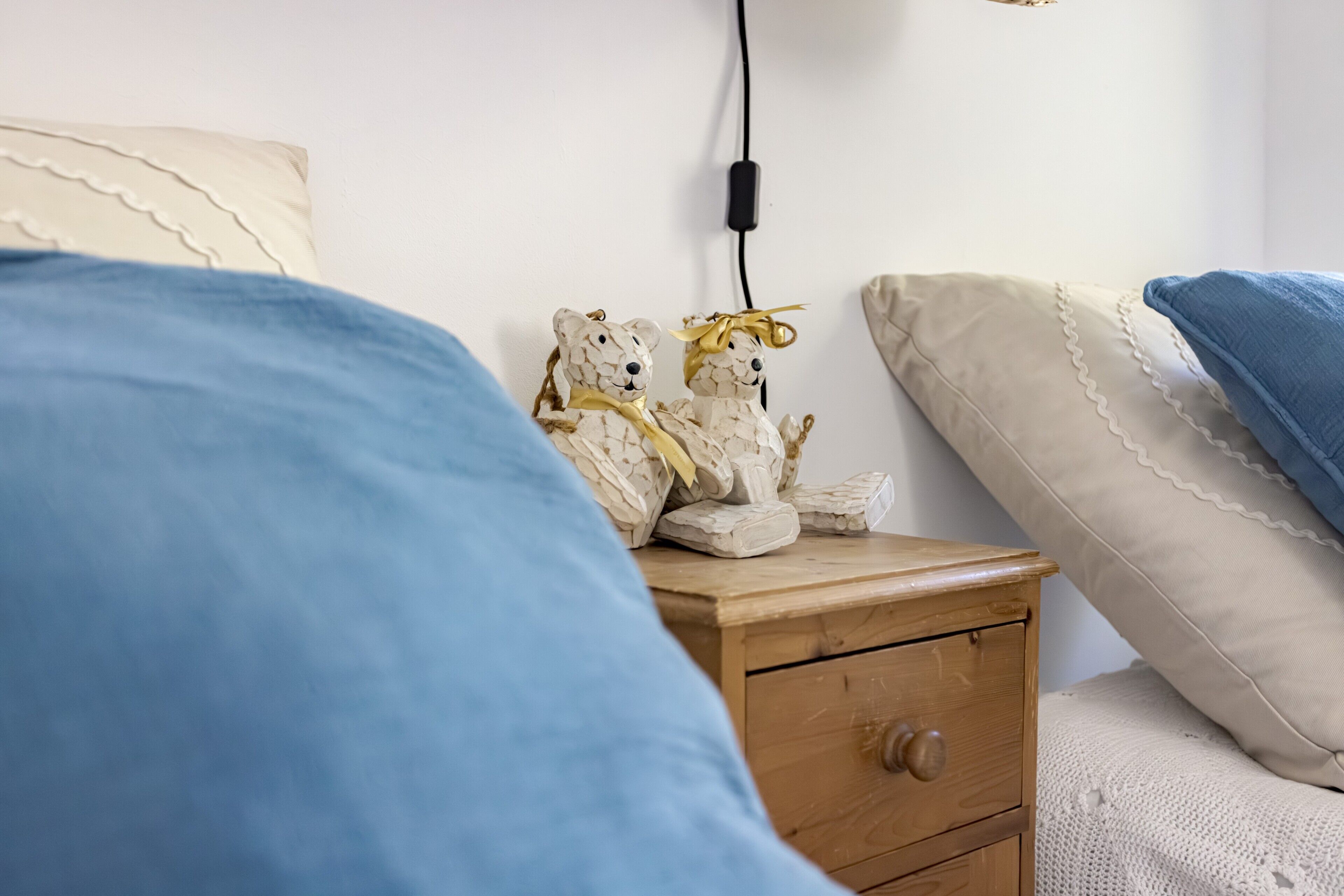 Un dormitorio con una mesita de noche de madera, una cama con manta azul, un almohadón blanco y un almohadón color beige. Hay un enchufe en la pared.