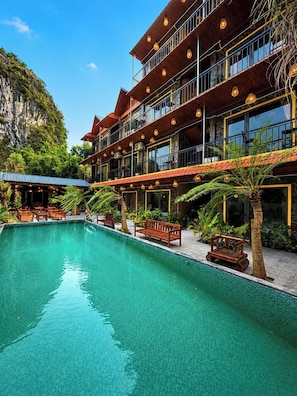 Un hotel de varios pisos con piscina, mobiliario exterior y abundante vegetación.