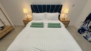 A neatly made bed with white bedding and green towels, a dark blue headboard with gold accents, and two bedside lamps on wooden nightstands. - Stylish 2 Bed Flat | Parking | Sutton Coldfield (Birmingham)