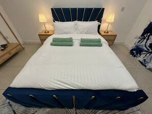 A neatly made bed with white bedding and green towels, a dark blue headboard with gold accents, and two bedside lamps on wooden nightstands.