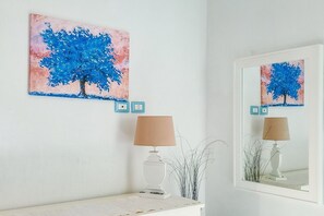 A room with a white wall, a blue and pink abstract painting, a lamp, and a mirror reflecting the painting. - Borgo Rio Favara 6 - Moon (Ispica)