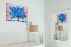 A room with a wall-mounted painting of a tree, a lamp, and a mirror reflecting the painting. - Borgo Rio Favara 6 Luna (Sicilia)