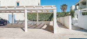 A covered outdoor area with a car parked under a white pergola, a paved walkway, and a building with a blue door in the background. - Borgo Rio Favara 6 Luna (Sicilia)