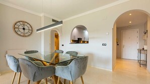 A dining area with a glass table and four chairs, a wall clock, and a kitchen visible through an archway. - 250m² PENTHOUSE DUPLEX- Marbella: 3 bedrooms, 3 bathrooms, solarium, close to the sea, golf courses (marbella)