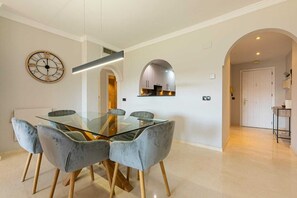 A dining area with a glass table and four chairs, a wall clock, and a kitchen visible through an archway. - 250m² PENTHOUSE DUPLEX- Marbella: 3 bedrooms, 3 bathrooms, solarium, close to the sea, golf courses (marbella)