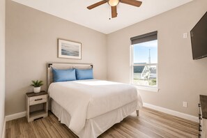 Ein Schlafzimmer mit Bett, Nachttisch, Topfpflanze, Deckenventilator, Fenster mit Blick und einem an der Wand montierten Fernseher.