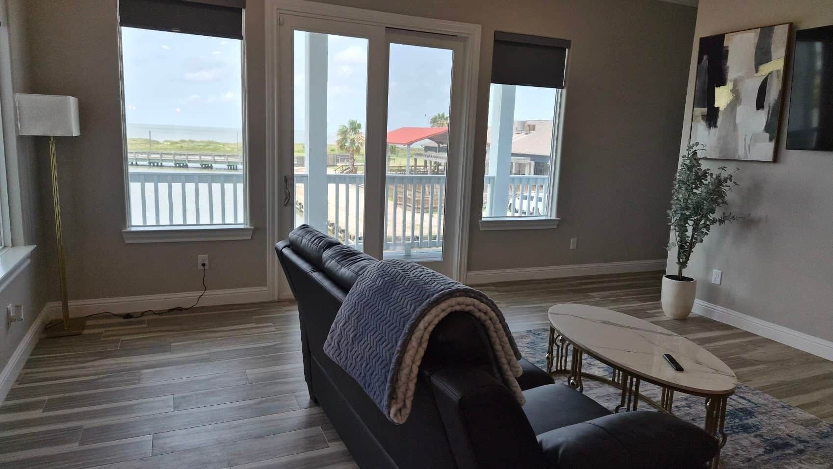 Een woonkamer met een donker leren bank, een klein rond tafeltje en uitzicht op het strand door grote ramen.