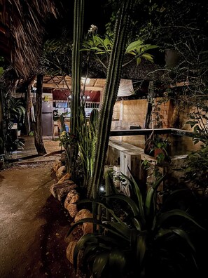 Un espace extérieur aménagé pour la nuit, doté d’une structure à toit de chaume, d’un réfrigérateur et de diverses plantes.