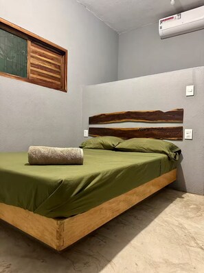 Uma cama de solteiro com colcha e almofadas verdes, cabeceira de madeira, janela com persianas e ar condicionado fixado na parede.