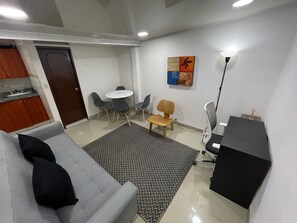 Uma sala de estar com sofá, mesa de jantar com cadeiras, uma cadeira de madeira pequena, escrivaninha com cadeira, um quadro na parede e um abajur.