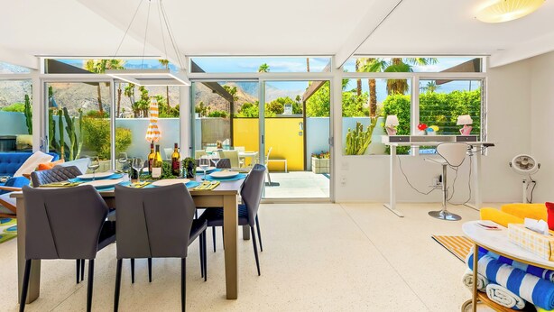 Ein moderner Essbereich mit Tisch für zwei, eine Bar mit Ventilator und Blick auf einen Garten durch große Fenster.