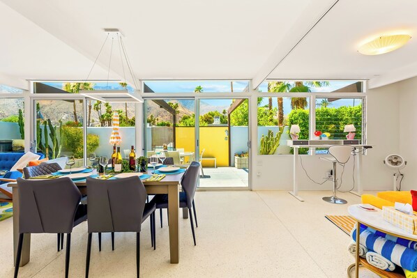 Ein moderner Essbereich mit Tisch für zwei, eine Bar mit Ventilator und Blick auf einen Garten durch große Fenster.
