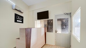 A reception area with a wooden reception desk, a mounted flat-screen TV, a door with a window, and a sign on the wall. - Classic Global Tower (Mathura)