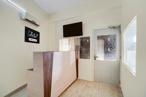 A reception area with a wooden reception desk, a mounted flat-screen TV, a door with a window, and a sign on the wall. - Classic Global Tower (Mathura)
