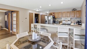 Una cocina moderna con armarios de madera, un refrigerador de acero inoxidable, un microondas y una cafetera. Hay una mesa de comedor de vidrio con sillas blancas.