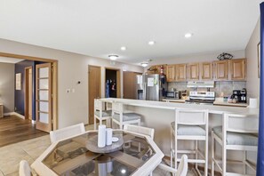 A modern kitchen with wooden cabinets, a stainless steel refrigerator, a microwave, and a coffee maker. There is a glass-top dining table with white chairs. - Parkside Escape|Seasonal Pool|Lake View|Pool View (Osage Beach)