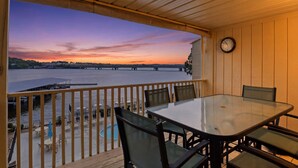 Un espacio exterior techado con una mesa y sillas, con vista a un cuerpo de agua y un horizonte urbano al atardecer.