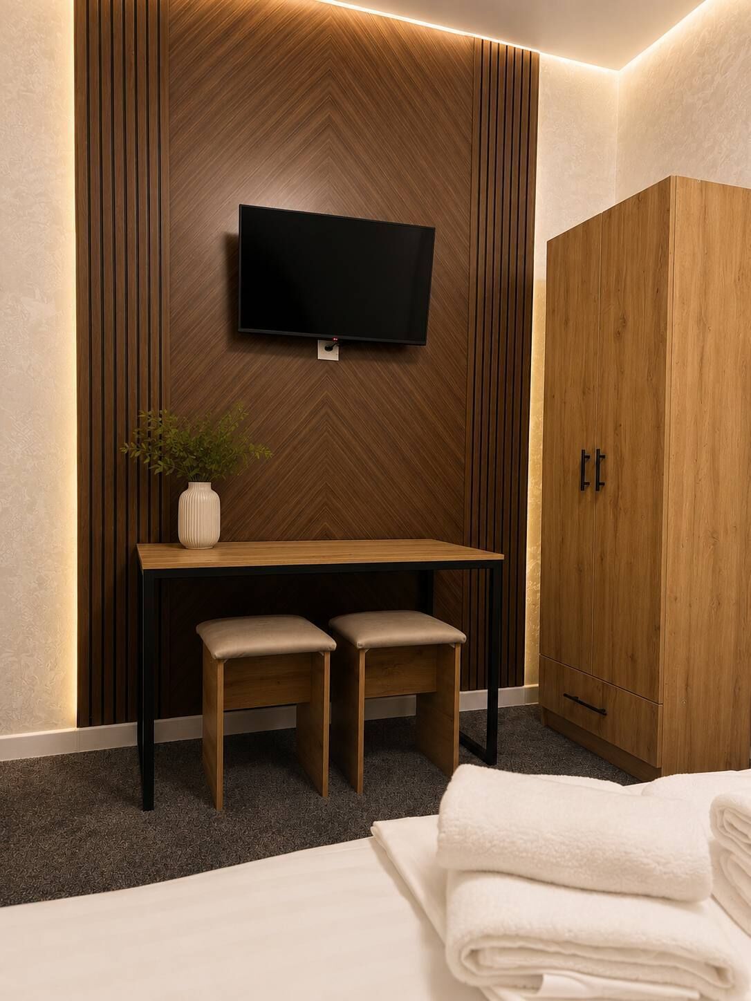 A hotel room with a wooden wall panel, a flat-screen TV mounted above a wooden desk with two chairs, a wardrobe, and a neatly folded white towel on the bed.