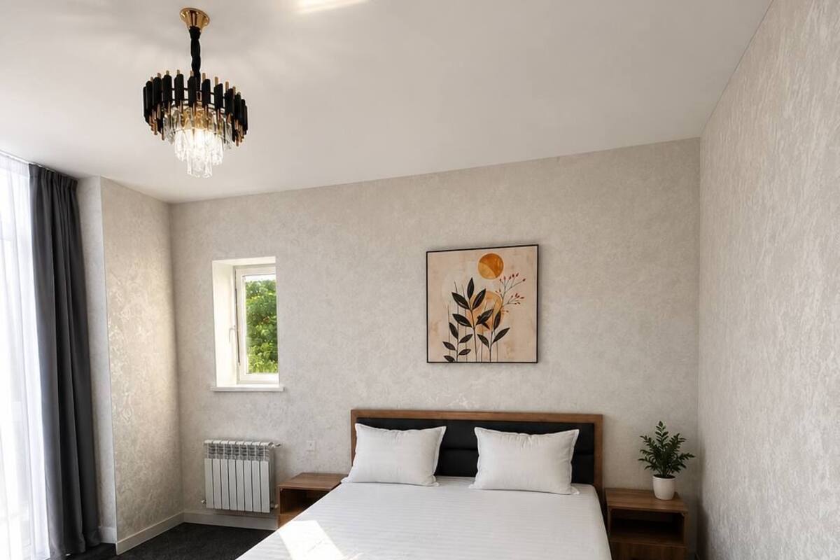 A hotel room with a large bed, a chandelier, a window, a radiator, and a plant.