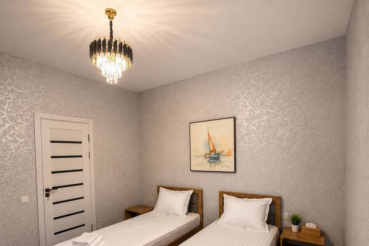 A bedroom with two beds, a chandelier, a painting, a door, and a nightstand.