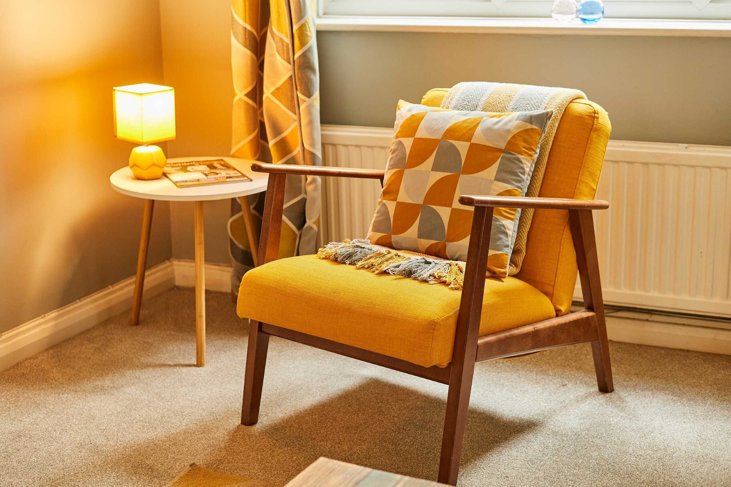 A yellow armchair with a checkered cushion, a small table with a lamp, and a window with curtains.