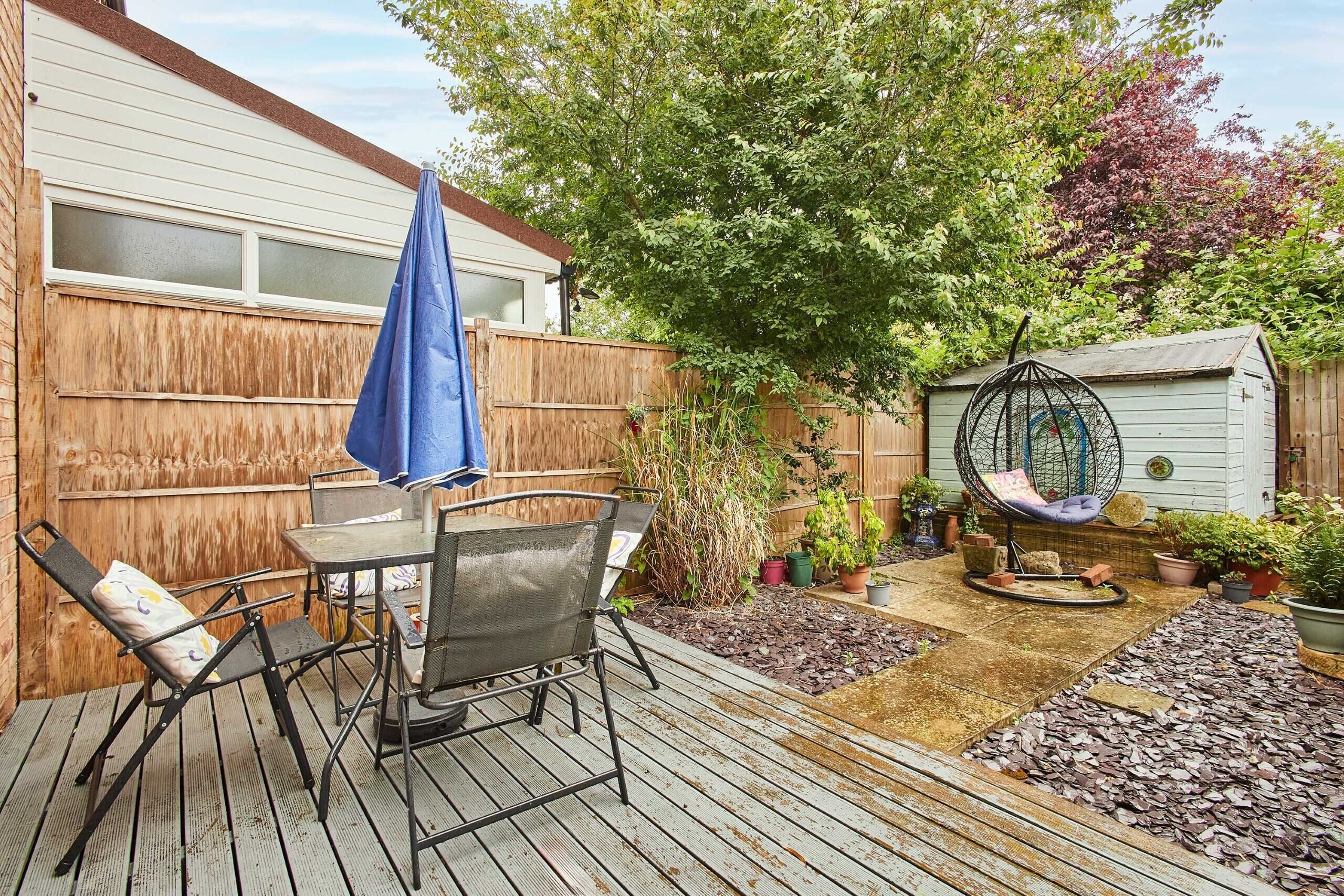 A wooden deck with a table and chairs, a hanging chair, and a garden area with potted plants and a shed.