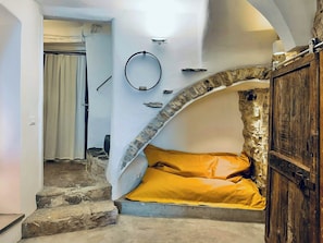 Ein Zimmer mit einem steinernen Bogen, der zu einem Bett mit gelber Matratze, einer Holztür und einer Steinwand führt.