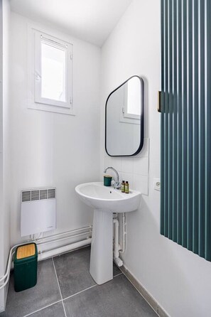 A bathroom with a white sink, a mirror, a window, a towel rack, and a green trash bin. - Renovated Studio 2 persons Nation Bastille Paris 11 (Paris)