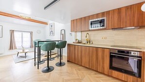 Una cocina moderna con armarios de madera, microondas integrado y horno. Hay dos taburetes verdes y una mesita redonda con un florero.