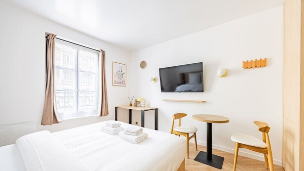 Habitación de hotel con cama, televisión, mesita, dos sillas y ventana con cortinas.