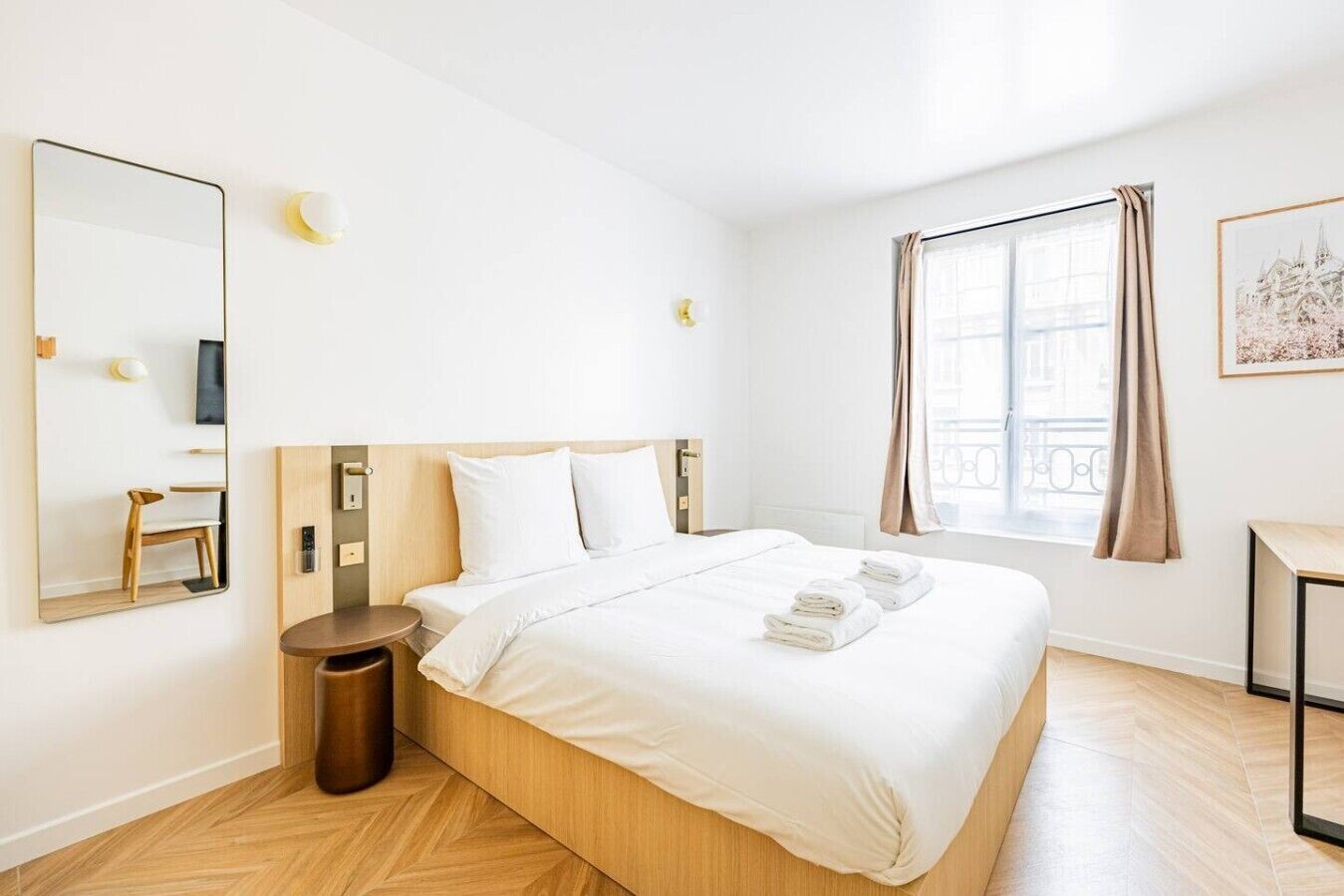 A hotel room with a bed, a round wooden nightstand, a mirror, a wall-mounted light, a window with curtains, and a framed picture on the wall.