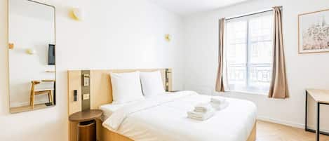 A hotel room with a bed, a round wooden nightstand, a mirror, a wall-mounted light, a window with curtains, and a framed picture on the wall.