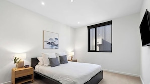 Ein modernes Schlafzimmer mit Bett, Nachttischen, einem an der Wand montierten Fernseher und einem Fenster mit Jalousien.