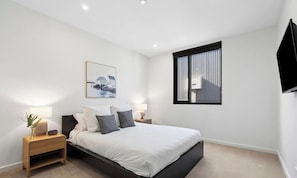A modern bedroom with a bed, bedside tables, a wall-mounted TV, and a window with blinds. - Bright 3-bed Apartment by Western Beach (Geelong)