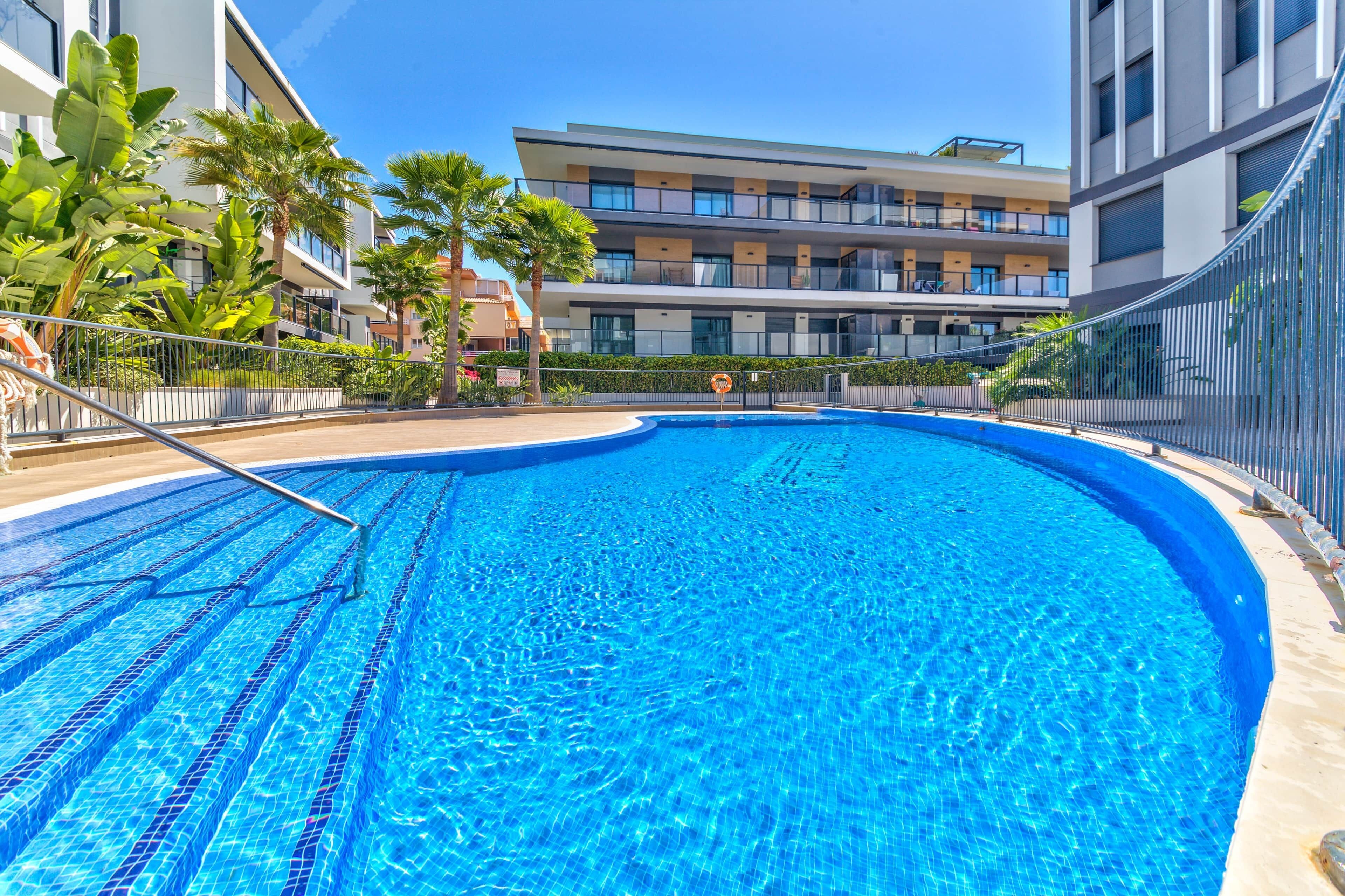 Una piscina con una escalera metálica, rodeada por una valla y contigua a un edificio de varios pisos con balcones.