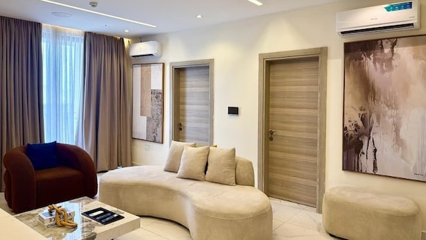 Ein modernes Wohnzimmer mit einer gebogenen Couch, einem kleinen Couchtisch und einem roten Sessel. Zwei Türen und eine Klimaanlage sind an der Wand angebracht.
