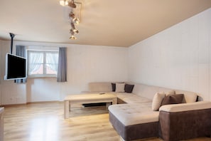 A modern living room with a sectional sofa, a flat-screen TV, a wooden coffee table, and a window with curtains.