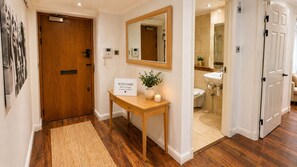 Une porte en bois, une pancarte de bienvenue, un miroir, une petite table avec une plante, et une salle de bain visible à travers une porte ouverte.