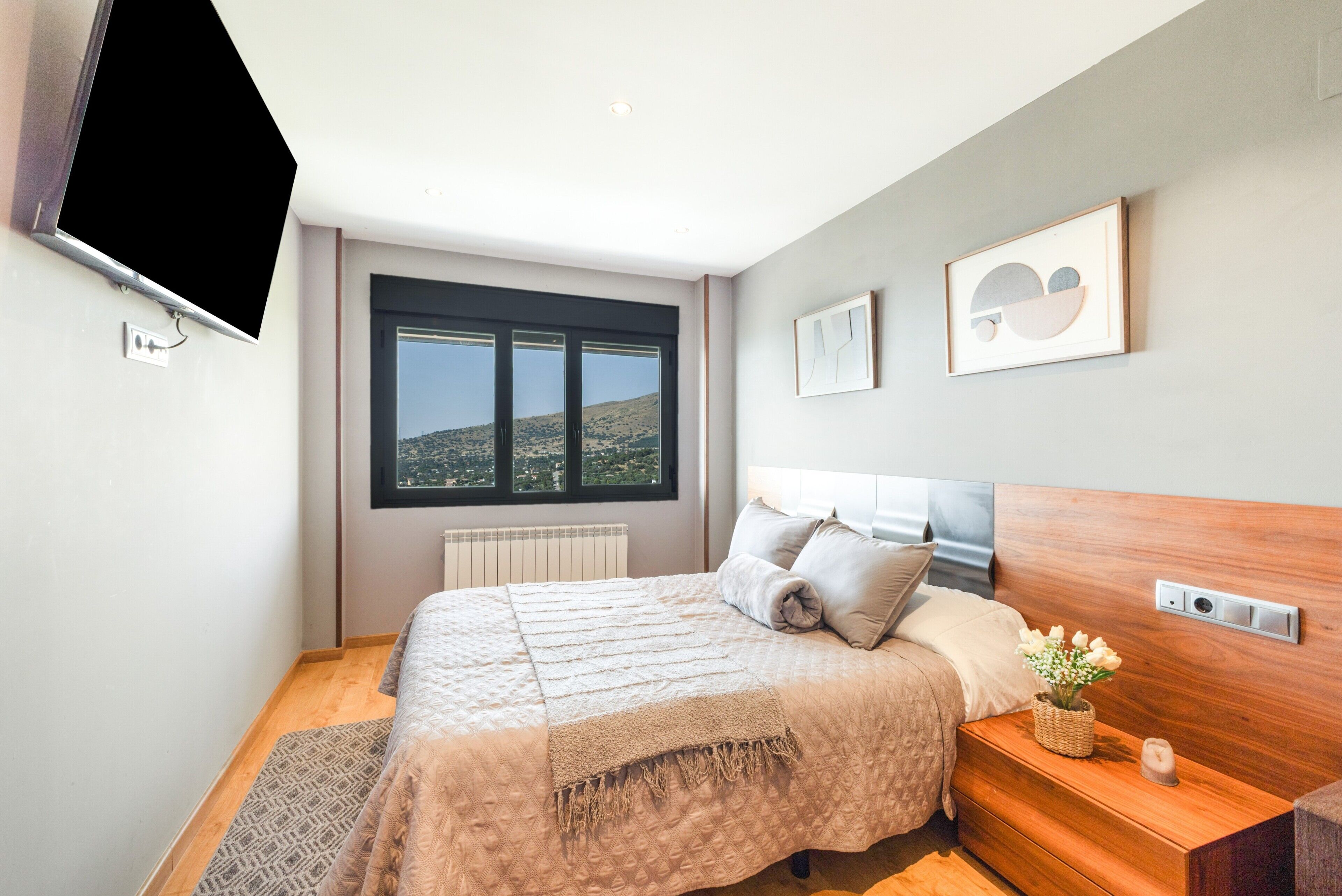 A modern bedroom with a large bed, a bedside table with a plant, and a view of the outdoors through a window.