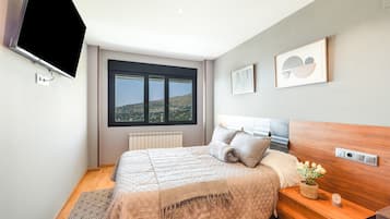 A modern bedroom with a large bed, a bedside table with a plant, and a view of the outdoors through a window.
