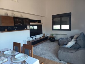A modern living space with a kitchen, dining area, and seating area, featuring a flat-screen TV, wooden cabinets, and a large window.