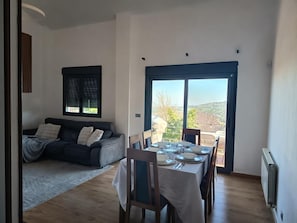 A living room with a sofa, a dining table set for two, and a large sliding glass door leading to a balcony with a view.