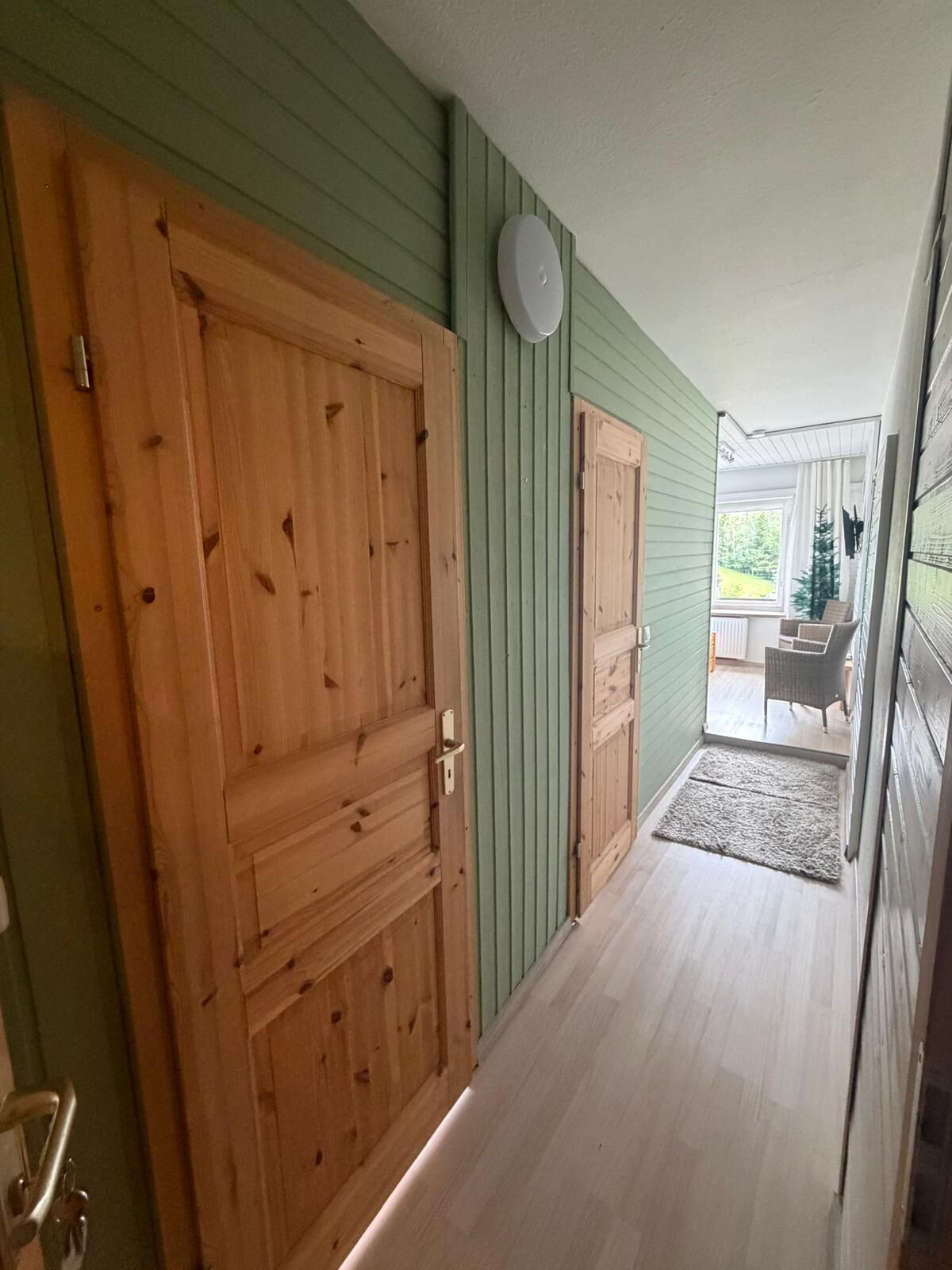 Un pasillo con puertas de madera, una pared verde y vistas a un espacio exterior con una silla y un árbol de Navidad.