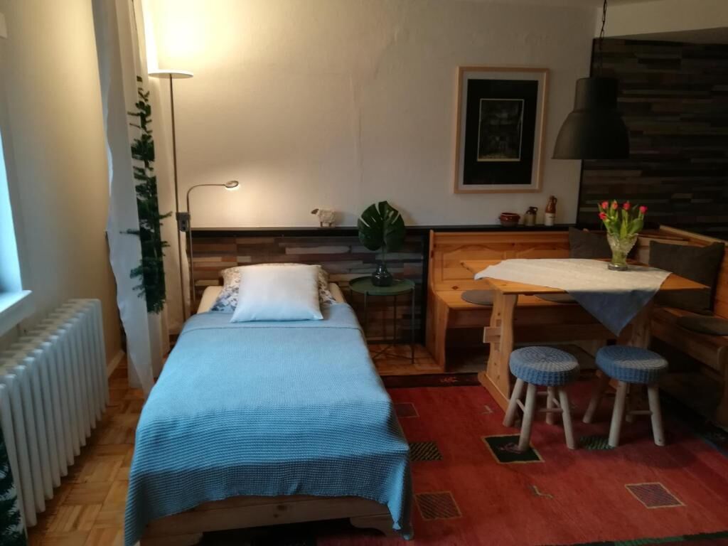 A small, cozy room with a bed, a desk, two stools, a radiator, a plant, and a framed picture on the wall.