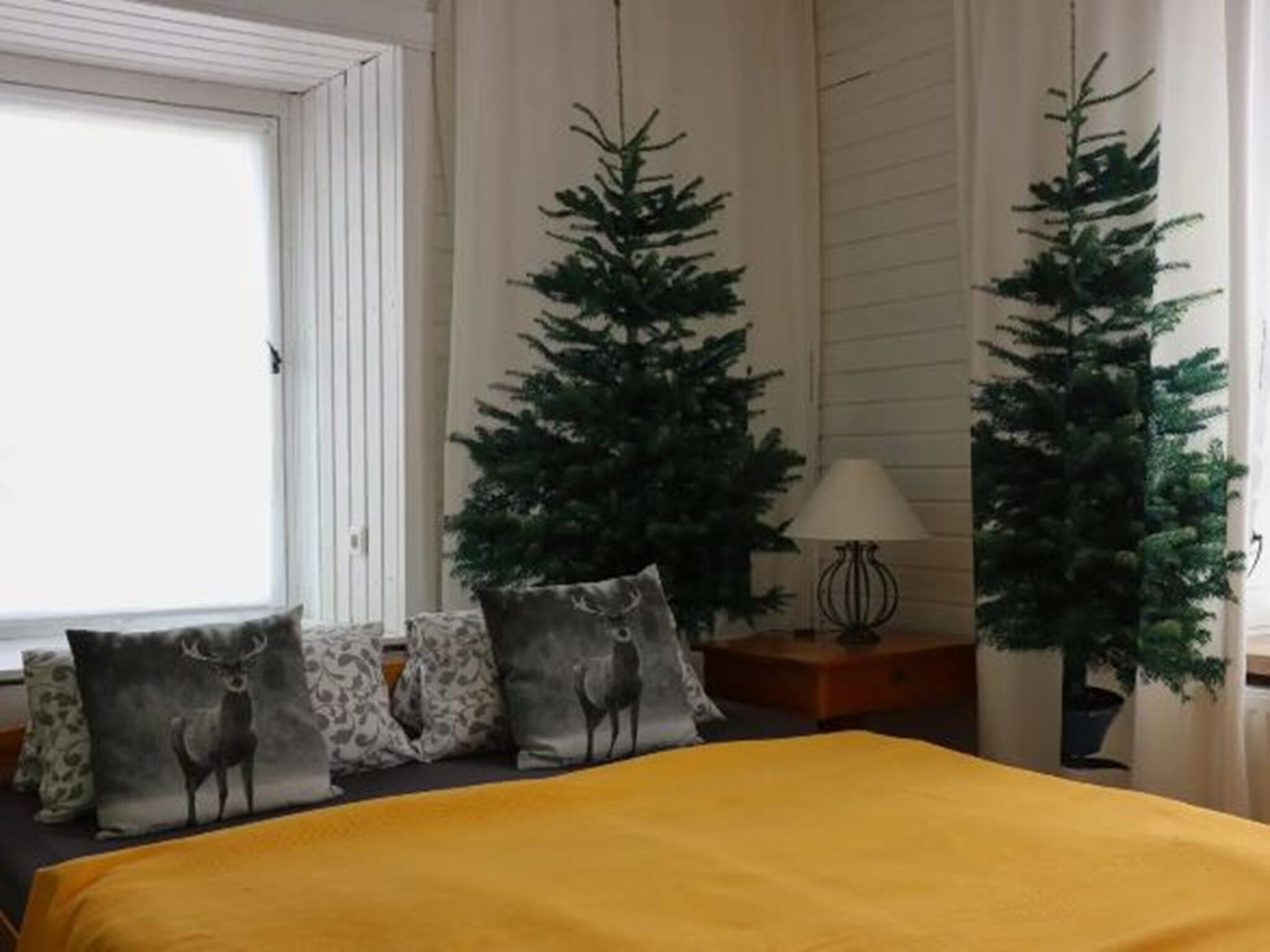 A room with a bed, a window, a lamp, and two artificial Christmas trees.
