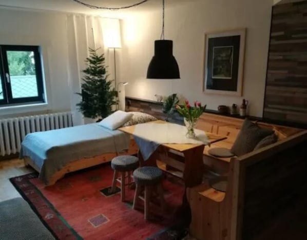 A compact room with a bed, a small table with stools, a radiator, a window, and a Christmas tree.