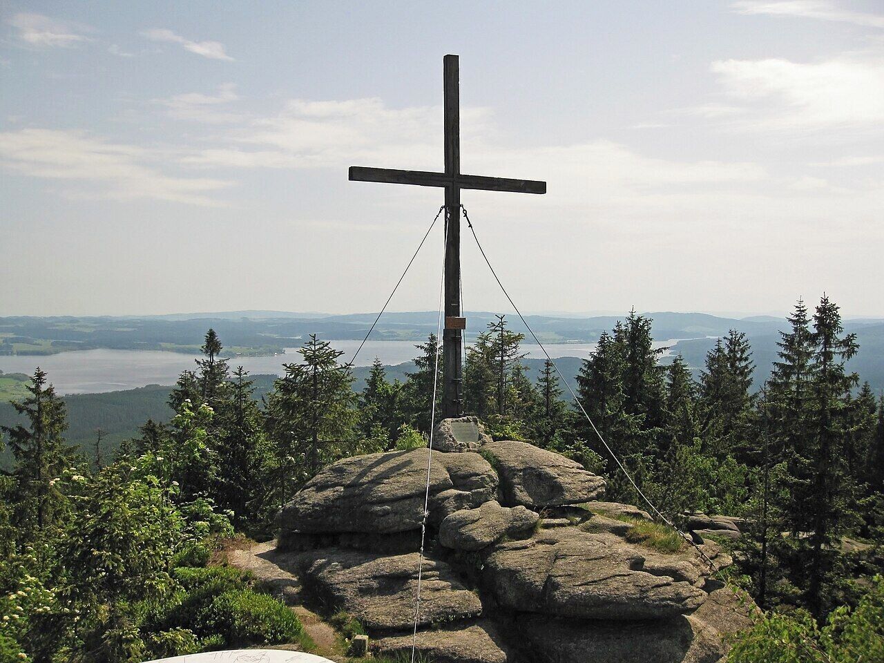 A cross on a rock with a view of a lake and mountains.