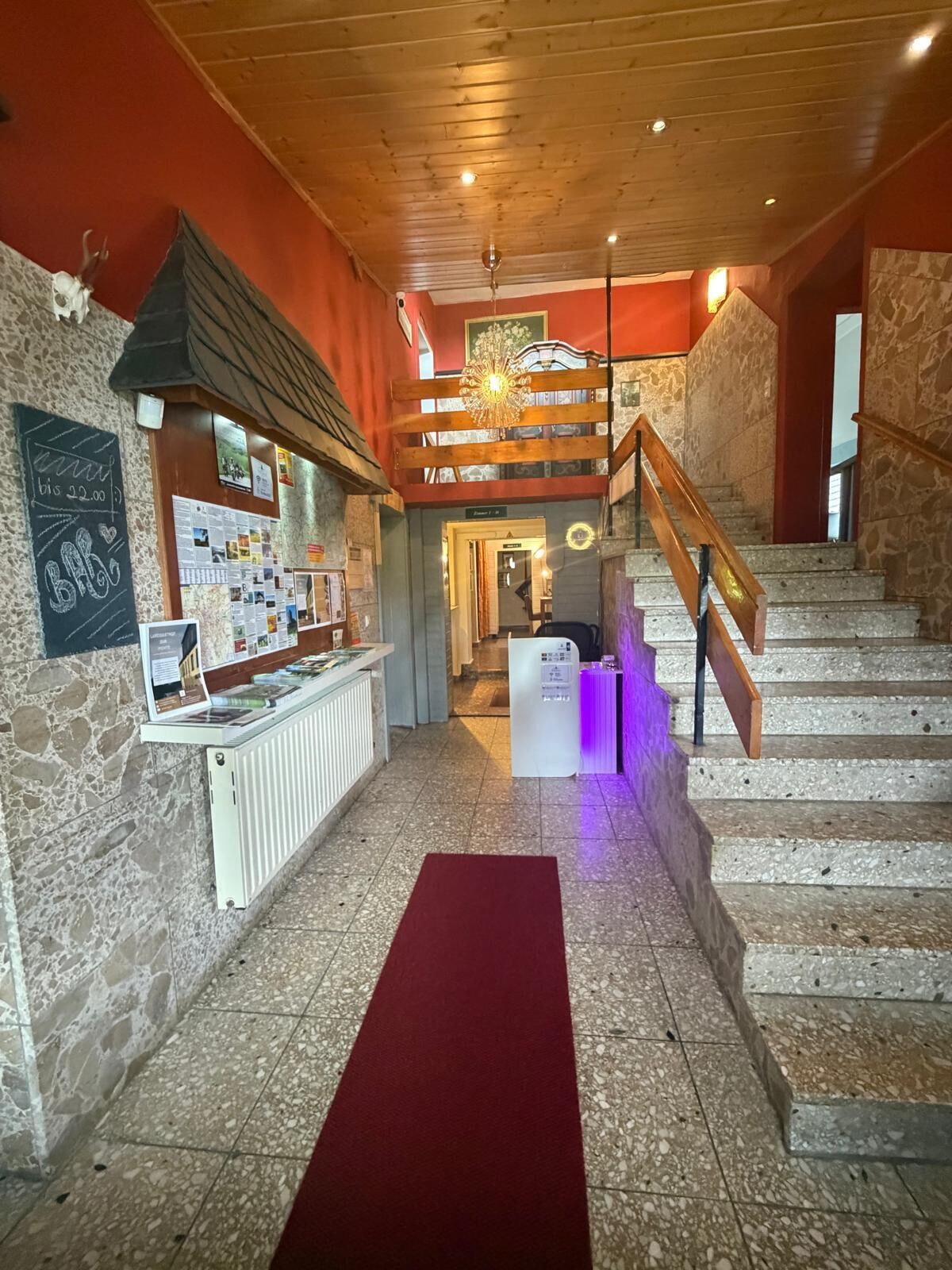 Un vestíbulo de hotel con mostrador de recepción, alfombra roja, una escalera y un candelabro.