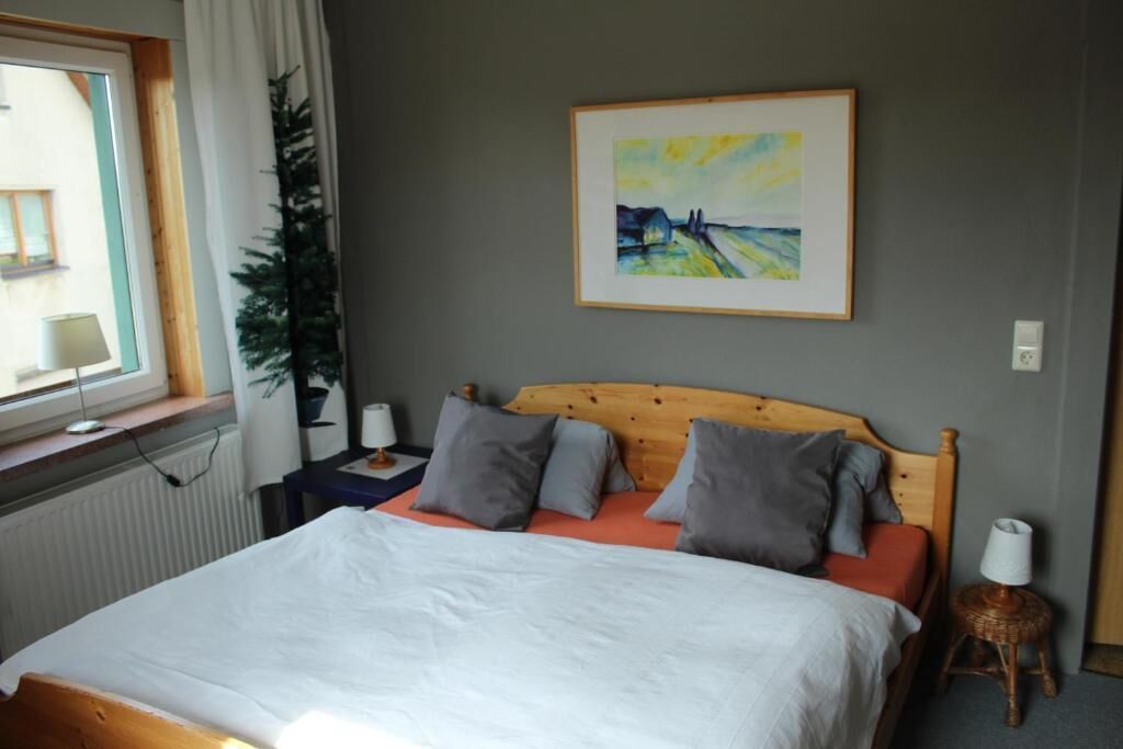 A bedroom with a bed, a nightstand, a lamp, a framed painting, and a window with curtains.