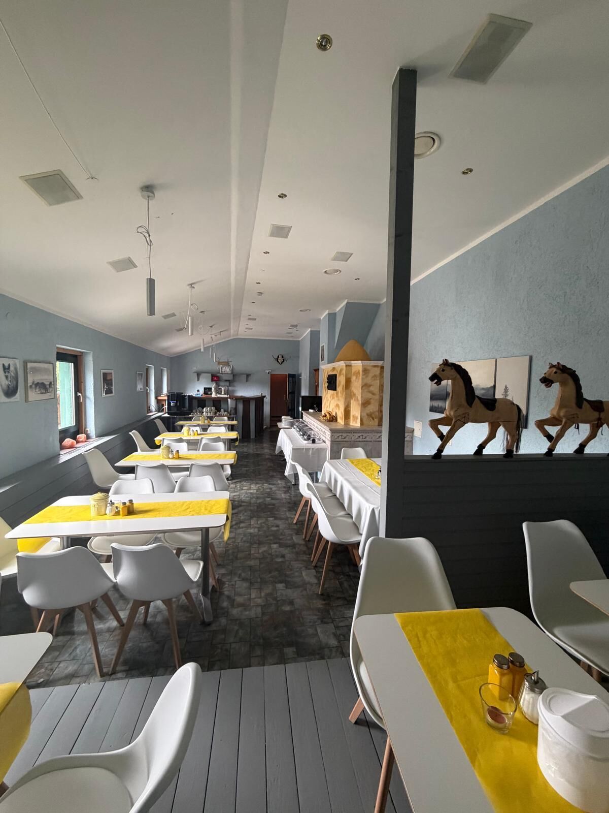 Un comedor con mesas y sillas, una cocina al fondo y estatuas decorativas de caballos en la pared.